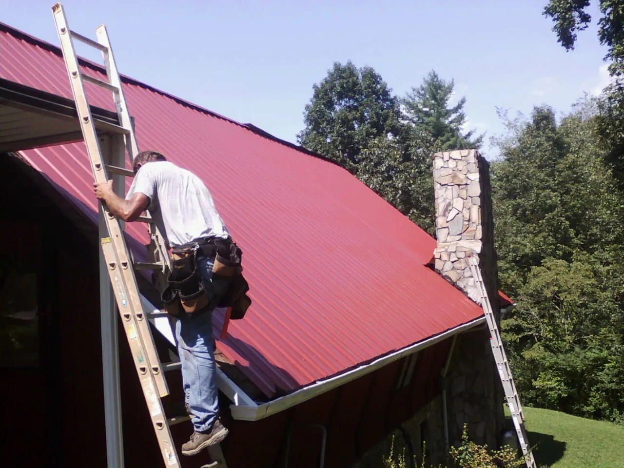 Roofer on a ladder beside a red metal roof and stone chimney — Soffit & Fascia Repair by Roofing Brothers in Twin Peaks